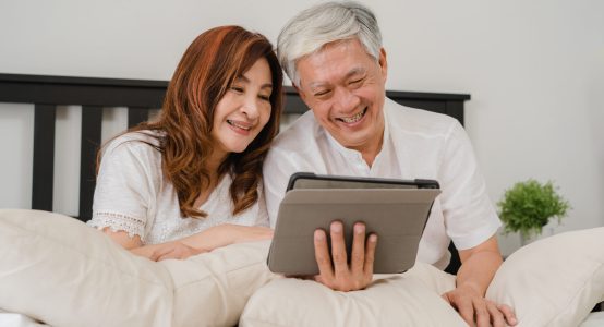 Asian senior couple using tablet at home. Asian Senior Chinese grandparents, husband and wife happy after wake up, watching movie lying on bed in bedroom at home in the morning concept.